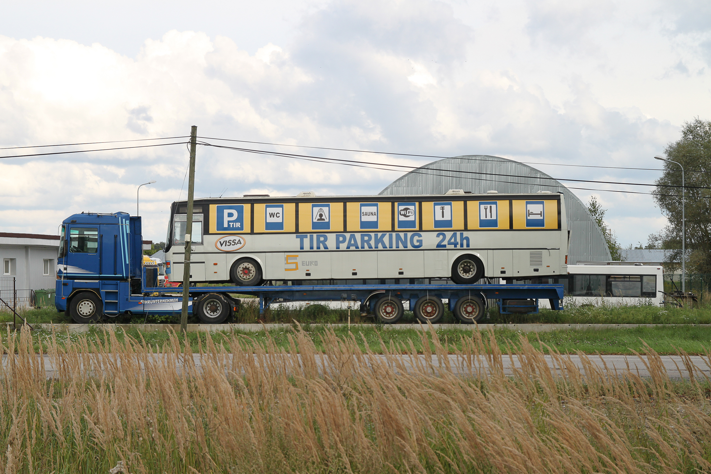 Corridor 1. E67 Adaži, Latvia: An old low-bed truck parked by the roadside carrying an even older municipal bus with large signs on the windows advertising the services of a rather informal-loo- king TIR truck drivers’ stop on a vacant, derelict industrial estate.